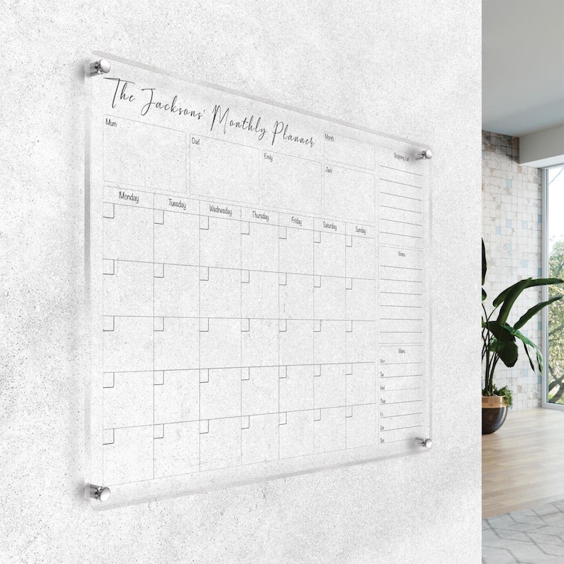 May include: Clear acrylic monthly planner with the text "The Jacksons' Monthly Planner" in elegant script. The planner features a grid layout for daily scheduling, with sections for notes and to-do lists. It is mounted on a textured white wall.