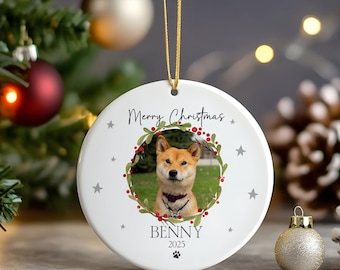 May include: White ceramic Christmas ornament with a photo of a Shiba Inu dog inside a wreath of greenery and red berries. The ornament has the text "Merry Christmas" and the dog's name, "Benny", along with the year 2025.