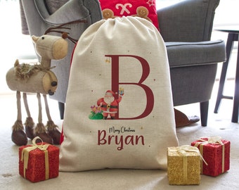 May include: A large beige Christmas gift bag with a red drawstring closure. The bag features a large burgundy letter "B" with a Santa Claus illustration and the words "Merry Christmas Bryan" in burgundy. Several wrapped gifts are in front of the bag.
