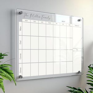 May include: A clear acrylic family planner with the text "The Miller Family" at the top. The calendar is divided into sections for each day of the week and includes spaces for family members' names. It is mounted on a wall with silver fixings.