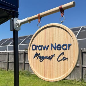 May include: A round wooden sign with the text Draw Near Magnet Co. in dark blue lettering. The sign hangs from a light-colored wooden rod by two brown leather straps with metal rings. The rod is attached to a white metal bracket on a black pole.