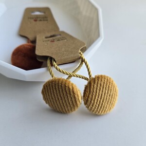 Könnte beinhalten: Zwei senffarbene Haargummis mit Cordstruktur und goldfarbenem Seil. Die Haargummis sind an braunen Anhängern mit dem Text "Handmade with love" befestigt. Die Haargummis befinden sich in einer weißen, geometrisch geformten Schale.