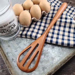 May include: A wooden kitchen utensil with a long handle and a slotted, paddle-shaped head. The tool features a smooth, natural brown wood grain finish and is positioned diagonally across a galvanized metal tray next to a glass sugar jar.