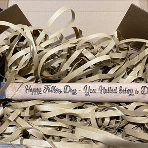 May include: A wooden hammer with a black metal head. The handle is engraved with the text "Happy Father's Day - You Nailed being a Dad". The hammer is surrounded by brown packing paper.