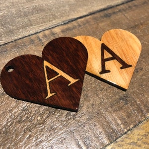 Puede incluir: Dos adornos de madera en forma de corazón con la letra "A" grabada en cada uno. Un corazón está teñido de un color marrón oscuro, mientras que el otro es de color madera natural.