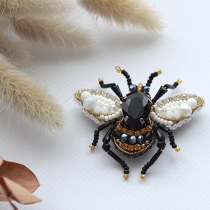 Puede incluir: Un broche de abeja negro y dorado con detalles de perlas blancas. La abeja tiene un cuerpo negro con detalles dorados y un cristal negro para la cabeza. Las alas están hechas de perlas blancas y cuentas negras.