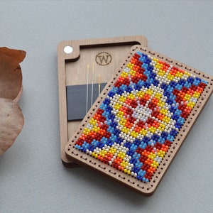 May include: Wooden needle case with a colorful beaded design. The case is open, revealing needles and a black magnetic holder. The beaded design features a geometric pattern in red, blue, yellow, and white. The case has a wooden frame with a stitched border.