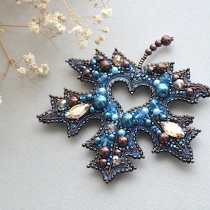 Può includere: Spilla a forma di foglia fatta a mano, con perline blu e marroni. La spilla presenta un ritaglio a forma di cuore ed è adornata con varie perline e cristalli. Il design della foglia è dettagliato con un'intricata lavorazione di perline.