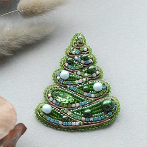 Könnte beinhalten: Handgefertigte Weihnachtsbaum-Brosche. Die Brosche besteht aus grünen Perlen und hat Akzente aus blauen und weißen Perlen sowie einen großen grünen Edelstein. Ein festliches Accessoire.