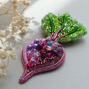May include: A beaded brooch in the shape of a beet with a green leafy top. The beet is made of pink, purple, and red beads, and the leaves are made of green beads. The brooch is a unique and whimsical piece of jewelry.