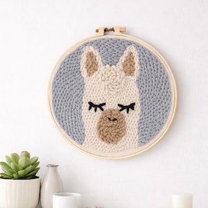 Puede incluir: Un tapiz de pared redondo y hecho a mano con un diseño de llama. La llama tiene una cara blanca, orejas y nariz marrones y pestañas negras. El fondo es de color gris claro y la pieza está enmarcada en un aro de madera.