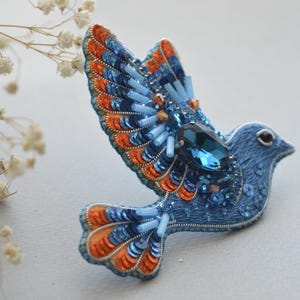 Può includere: Una spilla a forma di uccello blu e arancione con una grande pietra preziosa blu al centro. L'uccello è fatto di tessuto e decorato con paillettes e perline.