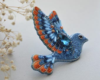 May include: A blue and orange bird brooch with a large blue gemstone in the center. The bird is made of fabric and decorated with sequins and beads.