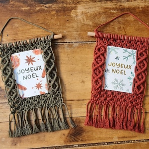 Puede incluir: Dos colgantes de pared de macramé con varillas de madera y lazos para colgar. Uno es verde oliva, el otro es de color óxido. Cada uno presenta una tarjeta con el texto "JOYEUX NOËL" e ilustraciones festivas.