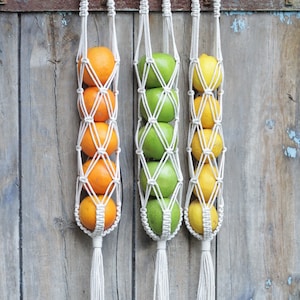 May include: Three macrame hanging fruit baskets with white cords and tassels. The baskets are filled with oranges, green apples, and lemons.
