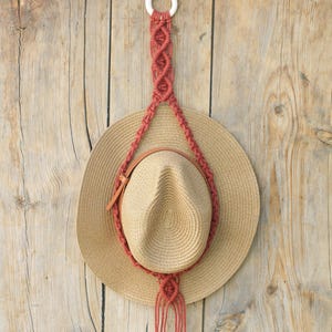 以下が含まれることがあります： 茶色の革バンドが付いた麦わら帽子が、マクラメの帽子ハンガーで吊り下げられています。 ハンガーは赤茶色のコードでできており、上部に白いリングが付いています。 背景は木のドアです。