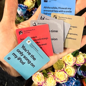 May include: A hand holding several small, colorful cards with song titles and lyrics, surrounded by a vibrant bouquet of roses in various colors. The cards feature song titles like "Good Days" and "You're the only song on my iPod."