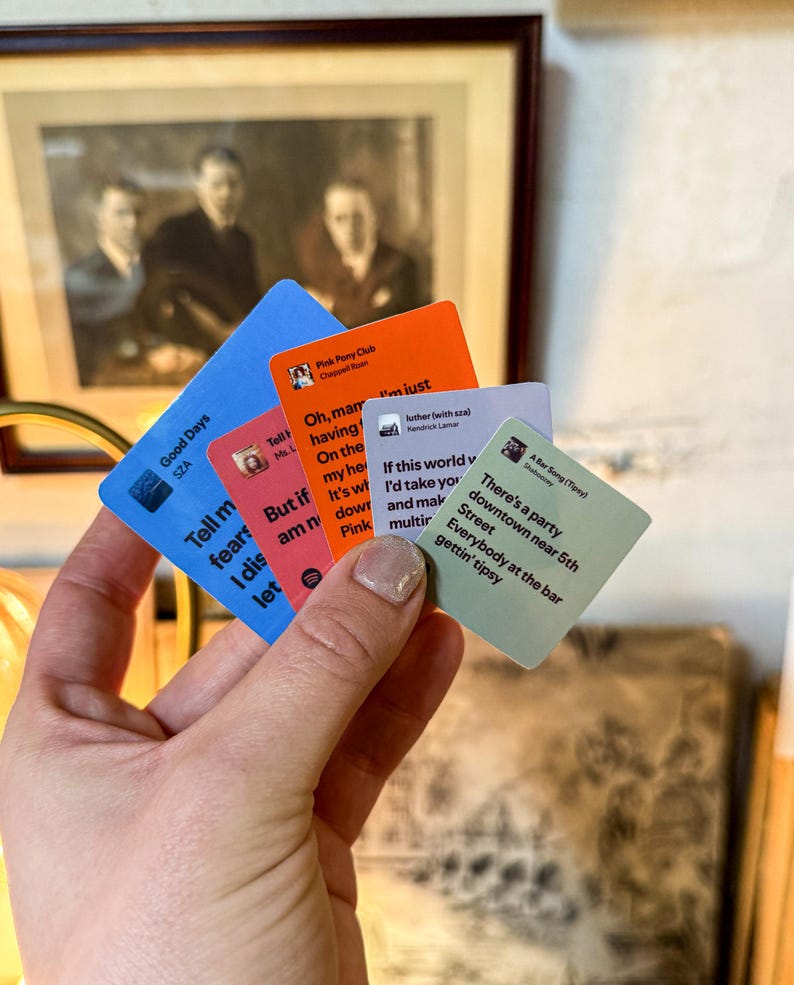 May include: A hand holding a stack of colorful square cards with song lyrics and artist names. The cards are blue, orange, red, and green. The text on the cards includes song titles and artist names such as "Good Days" by SZA.