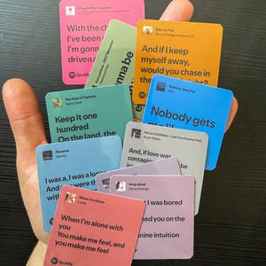 Op de afbeelding: Een hand die een verzameling vierkante kaarten vasthoudt met songteksten en artiestennamen. De kaarten zijn in verschillende kleuren, waaronder roze, oranje, blauw en groen. Het Spotify-logo is zichtbaar op sommige kaarten.