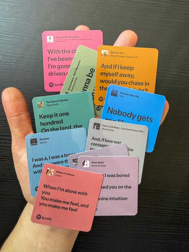 Op de afbeelding: Een hand die een verzameling vierkante kaarten vasthoudt met songteksten en artiestennamen. De kaarten zijn in verschillende kleuren, waaronder roze, oranje, blauw en groen. Het Spotify-logo is zichtbaar op sommige kaarten.