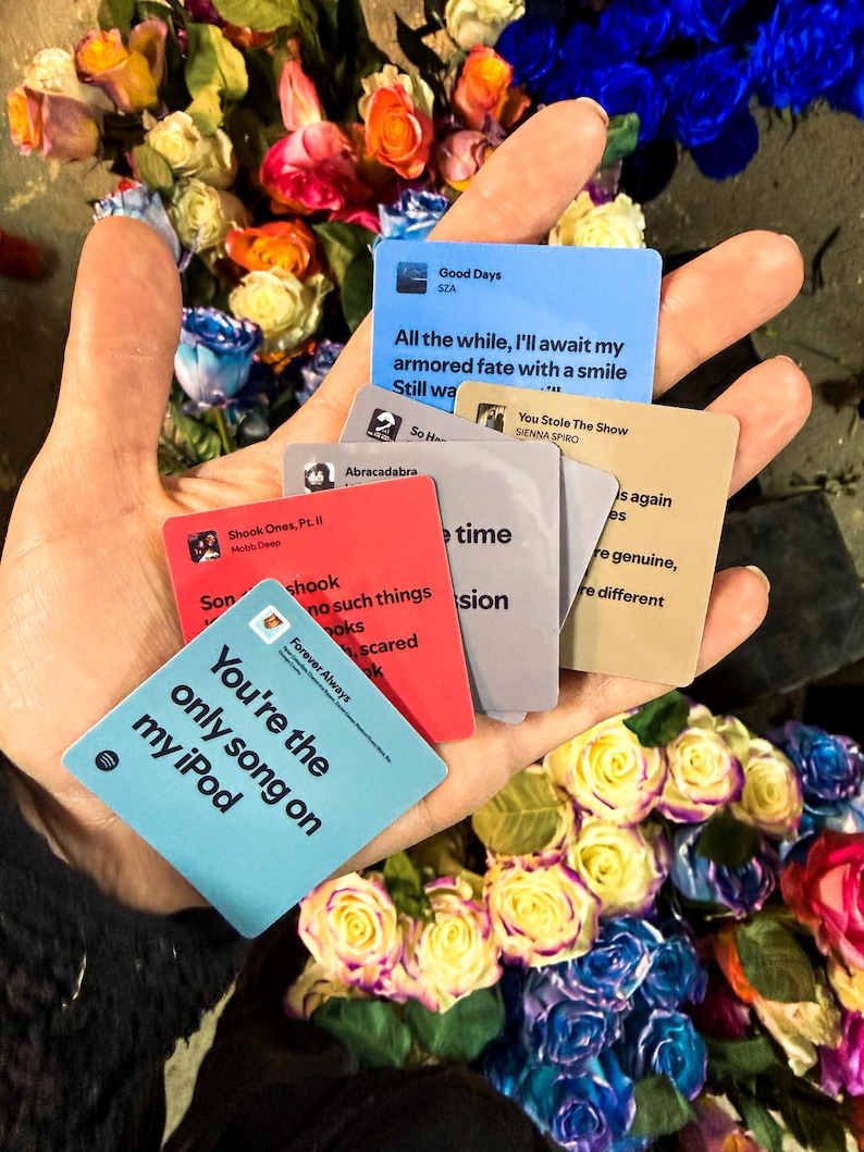 Op de afbeelding: Een hand die verschillende kleine kaarten met songtitels en songteksten vasthoudt, omringd door een kleurrijk boeket rozen. De kaarten zijn in verschillende kleuren, waaronder blauw, rood en bruin, met songtitels als "Good Days" en "You're the only song on my iPod."