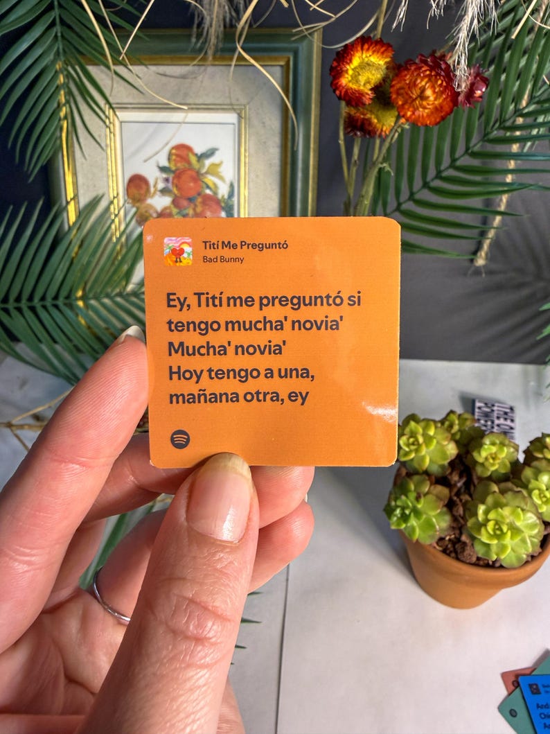 Op de afbeelding: Een oranje vierkante sticker met de tekst "Titi Me Pregunt&oacute;" van Bad Bunny. De sticker bevat Spaanse teksten en een Spotify-logo. De sticker wordt door een hand vastgehouden, met een vage achtergrond van planten en een ingelijste foto.
