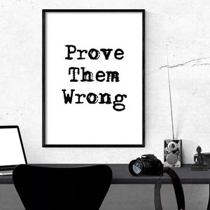 May include: A black and white framed print with the text "Prove Them Wrong" on a white background. The print is hanging on a white wall above a black desk with a laptop, a camera, a radio, and a small statue.