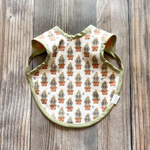 May include: A baby bib with a repeating pattern of cartoon cacti wearing glasses in brown pots on a white background. The bib has a green trim and straps, and is set against a wooden background.