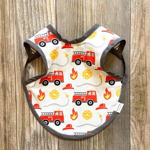 Peut inclure: Un bavoir blanc avec une bordure grise orné d'un motif de camions de pompiers rouges. Les camions de pompiers portent le numéro "No. 1". Le bavoir a une petite étiquette blanche avec le texte "Sweet Little Bean" dessus.