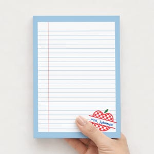 Peut inclure: Un bloc-notes bleu clair avec du papier ligné et une ligne de marge verticale rouge. Le coin inférieur droit présente un motif de pomme à carreaux rouges et blancs avec le texte "Mrs. Johnson" sur une bannière rouge diagonale.