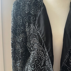 May include: A black beaded jacket with a V-neck and long sleeves. The jacket is decorated with a silver beaded floral pattern.
