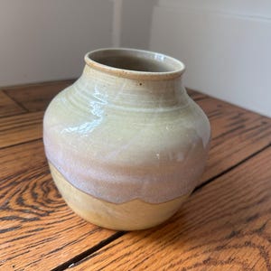 May include: A handcrafted ceramic vase with a rounded body and a narrow neck. The vase features a gradient of colors, transitioning from a light yellow base to a soft pink and cream upper section. The interior is a darker beige.