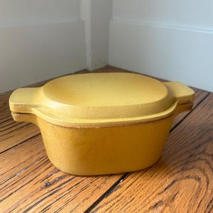 May include: A yellow casserole dish with a matching lid. The dish has two handles and sits on a wooden surface. The casserole dish is a warm, mustard yellow color.