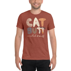 Puede incluir: Una camiseta de color óxido con un diseño gráfico de las palabras "CAT BUTT" en letras estilizadas de colores. El texto "scratch and sniff" está escrito en una fuente más pequeña debajo del texto principal.