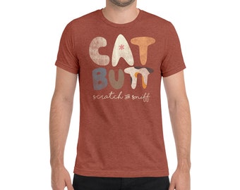 May include: A rust-colored t-shirt with a graphic design of the words "CAT BUTT" in colorful, stylized letters. The text "scratch and sniff" is written in a smaller font below the main text.