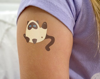 May include: A close-up of a person's arm with a temporary tattoo of a cartoon Siamese cat. The cat is brown and white with blue eyes and a long tail.