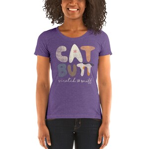 Puede incluir: Camiseta de manga corta de color morado con un diseño gráfico de las palabras "CAT BUTT" en letras coloridas y estilizadas. El texto "scratch & sniff" está escrito en cursiva debajo del gráfico.