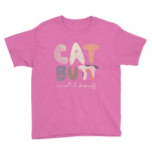 Puede incluir: Una camiseta rosa con las palabras "CAT BUTT" en letras redondeadas y coloridas. Debajo, "scratch & sniff" está escrito en una fuente manuscrita. La camiseta está hecha de un material suave.