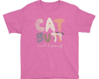 May include: A pink t-shirt with the words "CAT BUTT" in colorful, rounded letters. Below the text is "scratch & sniff" in a handwritten style font. The shirt is made of a soft material.