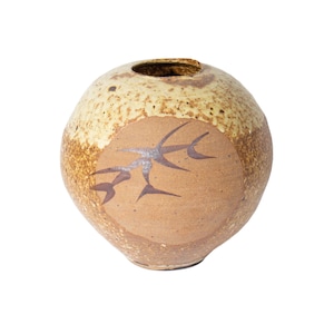 May include: A round, ceramic vase with a speckled, light tan upper half and a tan lower half. The lower half features a brown, stylized design. The vase has a small opening at the top.