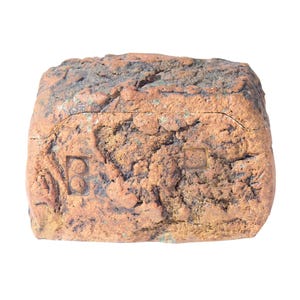 May include: A rectangular, weathered brick with a rough, textured surface. The brick is primarily reddish-brown with darker brown and black patches. The letters "B" and a square are imprinted on the surface. The brick is isolated on a white background.