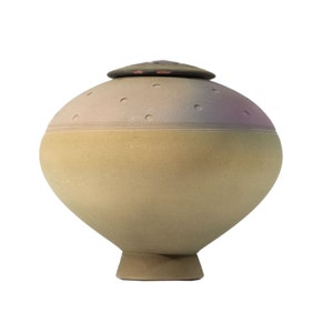 May include: A ceramic urn with a lid, featuring a speckled light brown and purple glaze. The urn has a rounded body and a small, round base.