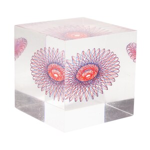 May include: A clear, cube-shaped decorative object with embedded red and blue spiral designs. The artwork is visible from all sides of the cube, creating a three-dimensional effect. The cube is set against a white background.