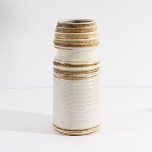 May include: A ceramic vase with a cylindrical body and a narrower neck. The vase is primarily off-white with horizontal bands of brown and tan. The texture appears to be ribbed, adding visual interest to the piece. The vase is likely for decorative use.