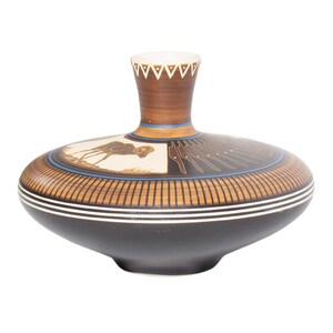 May include: A wide, low-profile ceramic vase with a narrow neck. The body features a dark brown base, a cream-colored panel with a brown ram illustration, and a series of vertical black lines. The neck has a white triangular pattern.