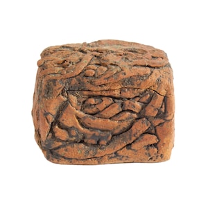 May include: A small, cube-shaped ceramic box with a textured, terracotta-colored surface. The exterior features raised, organic, root-like patterns in a darker brown tone, creating a rugged, uneven appearance across all visible sides of the object.