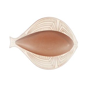 May include: A decorative, fish-shaped dish with a light beige, patterned border. The interior holds a smooth, oval-shaped object in a warm, earthy tone. The dish is set against a clean white background.