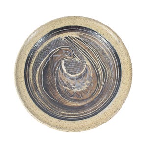 May include: A ceramic plate with a speckled beige glaze and a dark brown and blue design of a stylized bird in the center.
