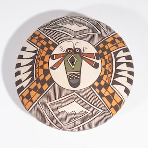 May include: A round, white ceramic plate with a geometric design in black, orange, and brown. The center of the plate features a stylized insect with green and black details.
