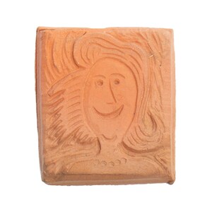 May include: A square terracotta stamp featuring a relief carving of a smiling face with long, flowing hair and a simple necklace. The surface is matte and earthy orange in color, showing a detailed portrait design pressed into the clay material.